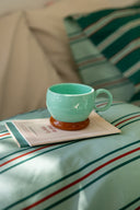 This image features a glazed ceramic coffee mug in a striking turquoise color with a brown base, displayed on a bed with striped bedding. The mug, handcrafted in Athens, Greece, showcases a unique design with a single rounded handle on each side and is adorned with the embossed Karavan logo at its base.